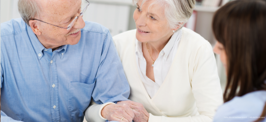 Älteres Ehepaar sitzt an einem Tisch und lächelt sich an, während es sich von einer jungen Frau beraten lässt.